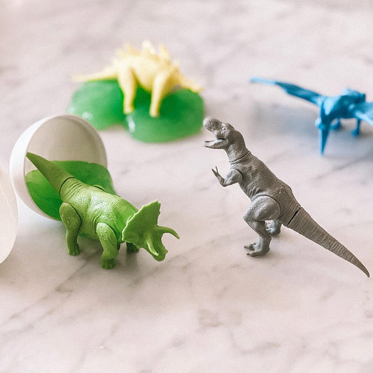 Image showing a T-Rex figurine and a Triceratops figurine on a table with two other figurines in the background and a cracked egg with green slime spilling out of it. 