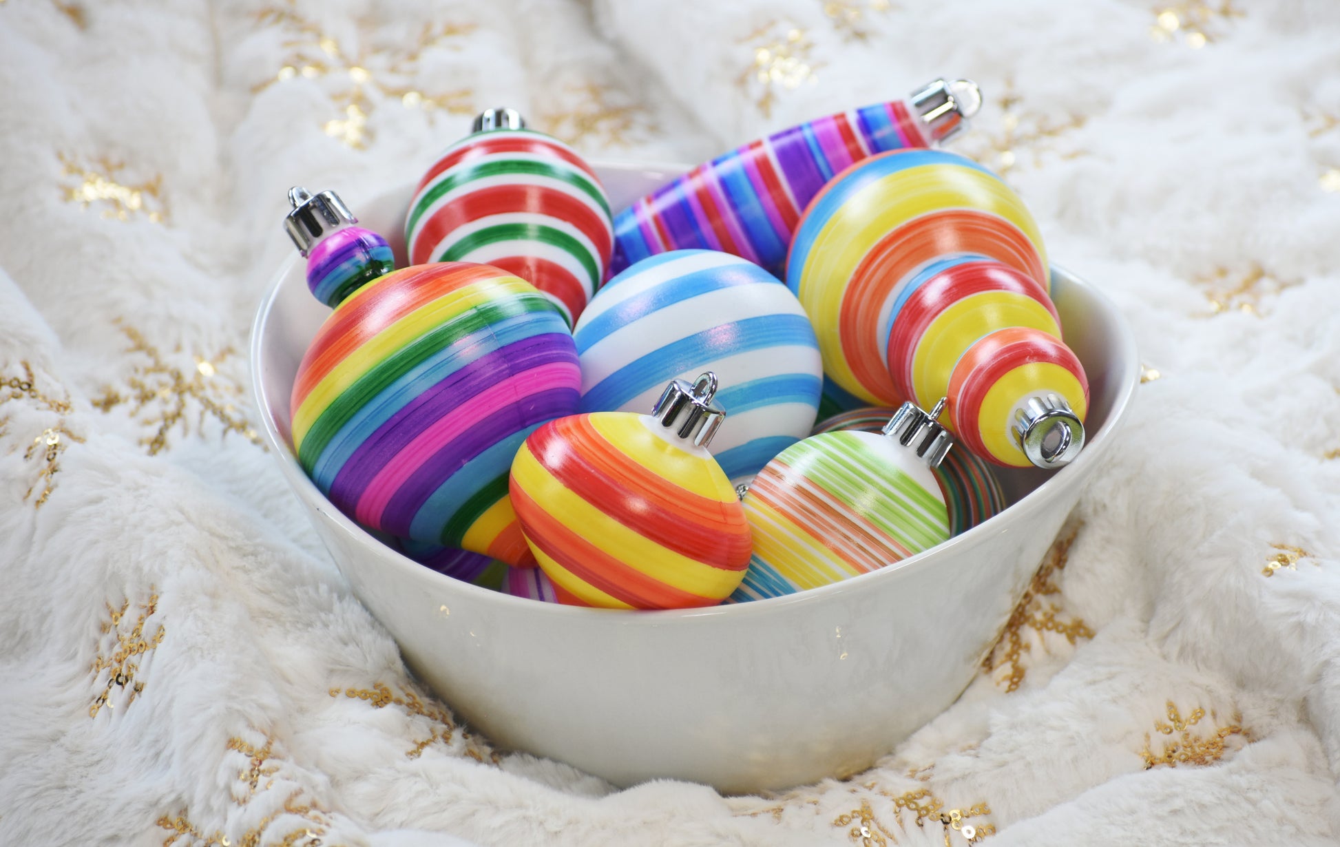 A white bowl with multiple colorful striped ornaments inside of it, sitting on a white and gold blanket. 