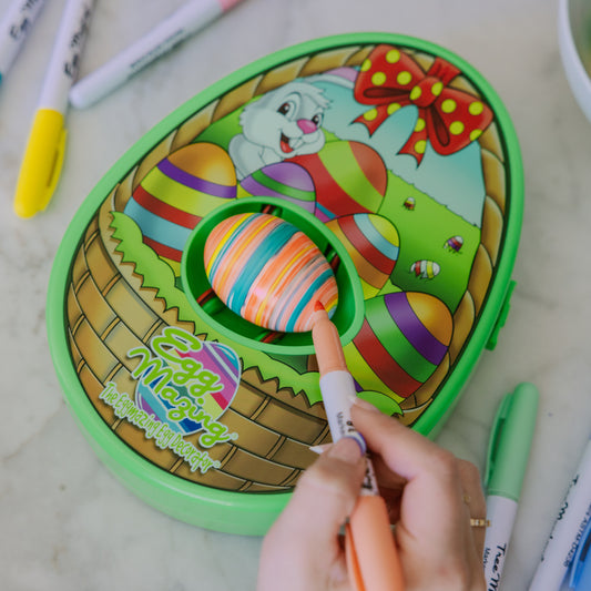 Image showing an egg shaped egg decorator on a table, with an egg in the machine and a hand holding a marker to the egg. 
